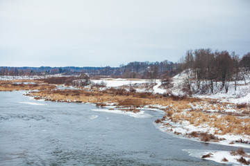 Winter landscape view of Venta river in Kuldiga, Latvia. Snow-covered stones and grass with ice edge in the turbulent flow. Last seasons brown grass and trees in the background.
