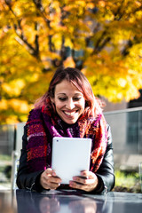 Fototapeta premium Portrait of a pretty young woman with a hipster style reading and laughing on her digital tablet computer sitting outside during a sunny autumn day. She's wearing a scarf