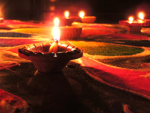 low angle shot of diwali oil clay lamps with intricate design burning with a glowing flame placed on multicoloured rangoli background