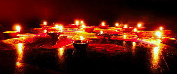 low angle view of round red blue green Diwali Rangoli pattern with dominant red orange drawn on dark black background illuminated by oil clay deep lamps