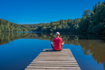 Fototapeta na wymiar Beulotte Saint Laurent, France - 09 12 2019: Hike in the circuit of the thousand ponds. a girl sitting on a wooden pontoon, doing yoga in front of the pond