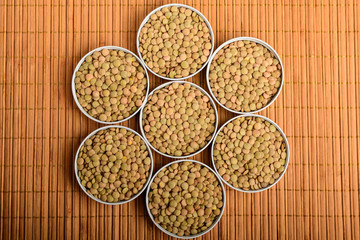 Seven bowls with organic fresh green lentil beans on a wooden table displayed as a flower in warm light, top view
