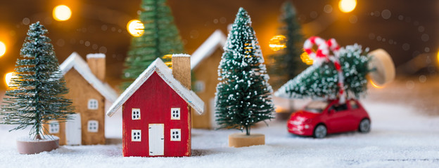 Miniature wooden houses and small red car with fir tree on the snow over blurred Christmas decoration background, banner