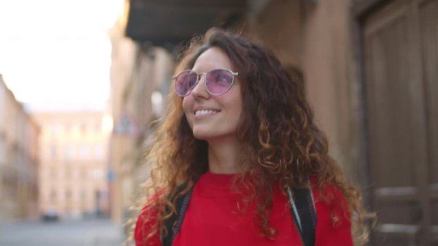 Tracking shot of happy curly woman in sunglasses holding mobile phone and walking along street in city. She is smiling and looking around