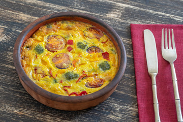 Ceramic bowl with vegetable frittata, simple vegetarian food. Frittata with egg, tomato, pepper, onion, broccoli and cheese on wooden table. Italian egg omelette