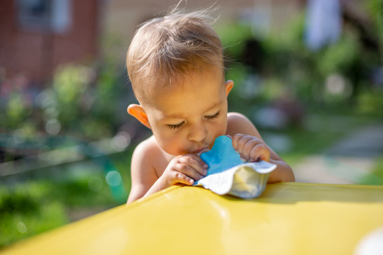 Baby Eating Fruit Puree In Pouch And Looking Into The Food In Front Of The Yellow Table. On The Background Is A Green Garden On A Sunny Day In Blur