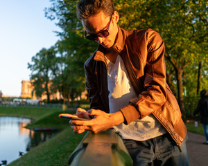 Obraz premium guy is looking at a smartphone. in the park in the light of the setting sun in sunglasses