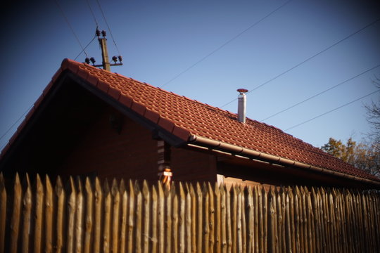 Wooden house with red tiled roof and blue sky. Fence made of sharp wooden stakes. Blurred.
