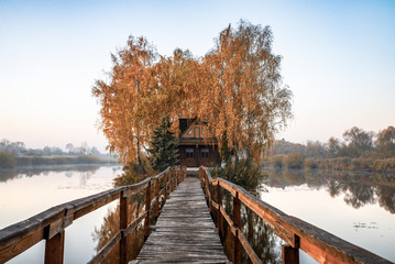 Morning landscape of a small house on an artificial island with a long wooden bridge. Around the house is a small lake with light fog. Autumn landscape