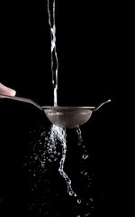 A stream of water pours through a small steel sieve in the hands of a man on a black background....