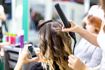 Naklejka premium close up stylist makes a girl client styling hair waving with curling irons in a hairdresser. customer girl looks into smartphone in blur. close up, soft focus