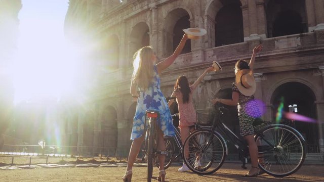 Three happy young women friends tourists with bikes waving hats at Colosseum in Rome, Italy at sunrise.