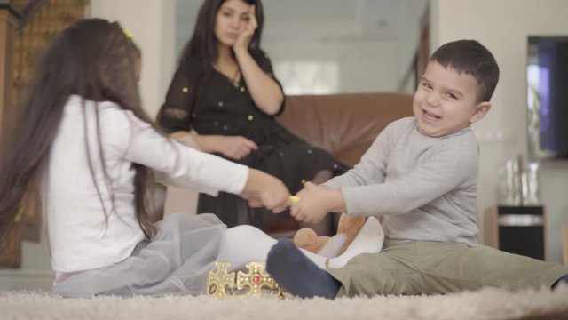 Two little Middle Eastern children fighting for the toy at the foreground and their mother holding her head desperately at the background. Siblings playing together at their cozy home.