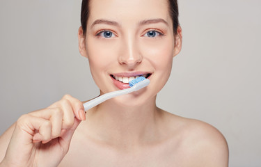 Young pretty girl with a toothbrush in her hands nearly her mouth with white teeth, with brown hair with clean fresh skin posing on a gray studio background