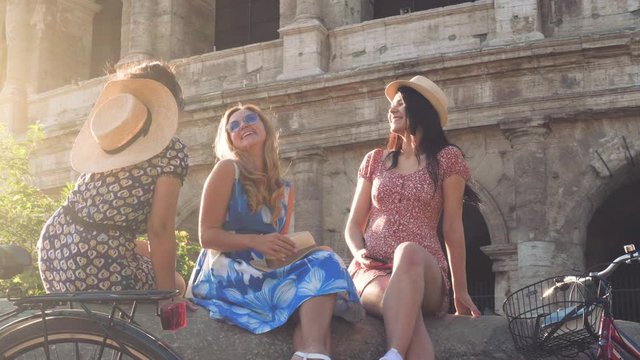 Three happy young women friends tourists with bikes having fun sitting on a column at Colosseum in Rome, Italy at sunrise.