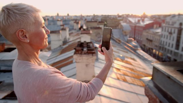 Medium shot of middle-aged woman with short blonde hair standing on old roof in downtown and making photos using smartphone