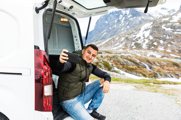 Naklejka premium Young man sitting in trunk car taking selfie on the phone in the mountains