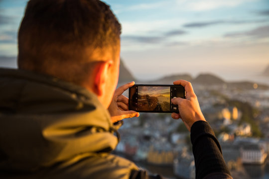 Young handsome man take photo on the phone of beautiful panorama of the city Alesund