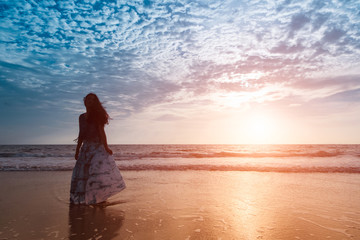 Fototapeta premium Asian girl wear Indigo dyed cotton long skirt,smiling and flower tucked behind her ear on the beach,blue sky and sea,orange light flare.
