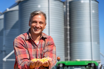 Smiling Farmer Showing Freshly Harvested Corn Maize Grains in front of Farm Grain Bins