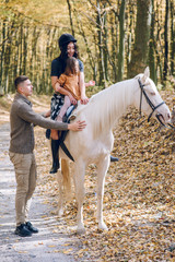 Obraz premium Young family is learning to ride a beautiful white horse in the autumn forest. Riding lesson