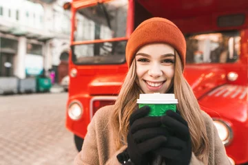 Poster Closeup portrait of happy girl with cup of coffee standing against red bus background, looking into camera and smiling. Street photo of cheerful girl on a walk in winter, warmed by warm coffee. © bodnarphoto