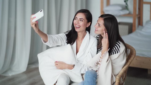 Two girls take selfie on phone, posing, show gesture of peace, smiling at home