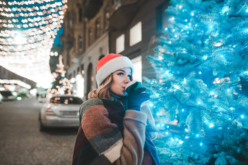 Portrait of a beautiful girl in a hat of Santa Claus stands on a Christmas-decorated street near a...