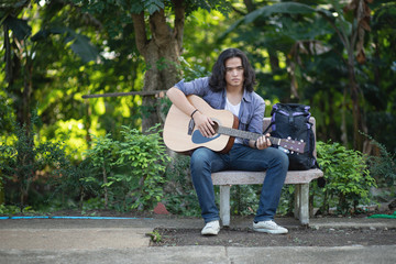 Asian long haired man is playing the guitar while he is traveling. With a rustic background and trees Under the soft sunlight in the day