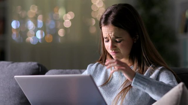 Sad woman reading bad news on laptop sitting on a couch in the night at home