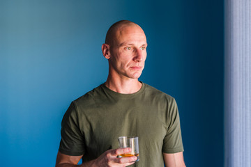 Portrait of young handsome man with short hair holding a glass of whiskey or brandy alcohol drink standing in front of blue wall looking trough the window