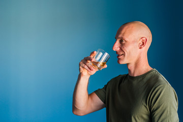 Portrait of young handsome man with short hair holding a glass of whiskey or brandy alcohol drink standing in front of blue wall side view profile