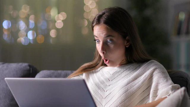 Surprised woman finding amazing on line news on laptop sititng on a couch in the night 