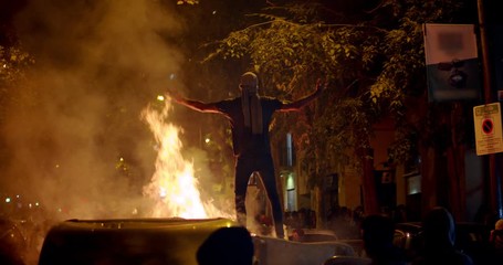 Young rebel riot revolutionary anarchist stands on trash and fire showing fuck as protest for independence on demonstration about the proces and political prisoners Demonstrator burns garbage bins in