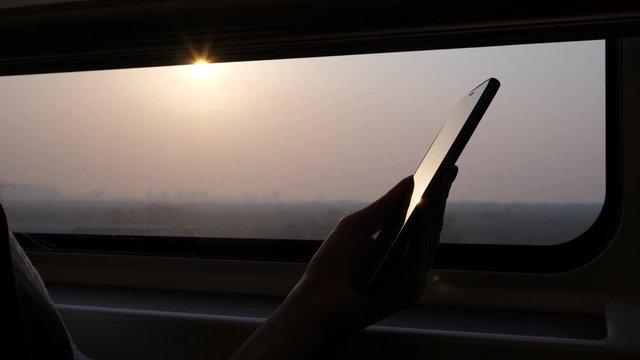 Passenger hold phone in hands, closeup high contrast shot against window, sun light shine and reflex at glossy screen of smartphone. Blurred background, modern high speed train ride to Beijing