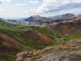 Reykjadalur valley with hot springs river with lush green grass meadow and hills with geothermal steam. South Iceland near Hveragerdi city. Summer sunny morning, blue sky.
