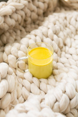 Golden milk in clear glass mug on chunky knitted blanket, turmeric oat milk, healthy supplement drink, copy space