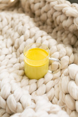 Golden milk in clear glass mug on chunky knitted blanket, turmeric oat milk, healthy supplement drink, copy space