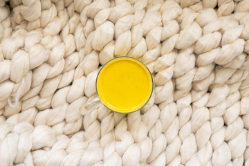 Golden milk in clear glass mug on chunky knitted blanket, turmeric oat milk, healthy supplement drink, copy space