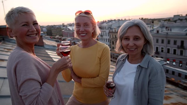 Medium shot of three middle-aged diverse women standing on vintage roof on sunset, drinking red wine, communicating and then looking at camera