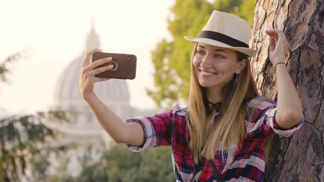 Happy young blonde woman with long hair and blue eyes laining on a tree, taking selfies with smartphone in front of Rome cityscape. Saint Peter church from Gianicolo panoramic view.