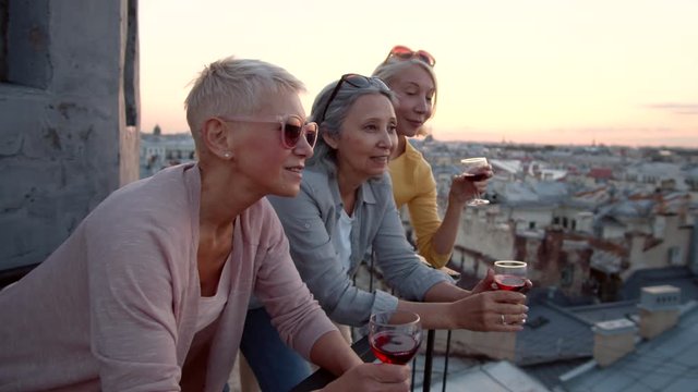 Medium shot of three middle-aged women standing on roof leaning on railing, holding glasses of red wine in their hands, smiling and discussing view