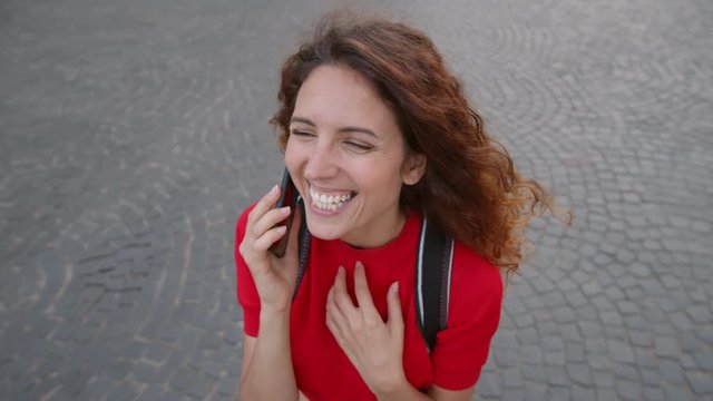 High angle tracking shot of ecstatic young woman with backpack laughing and talking on phone while looking around outdoors