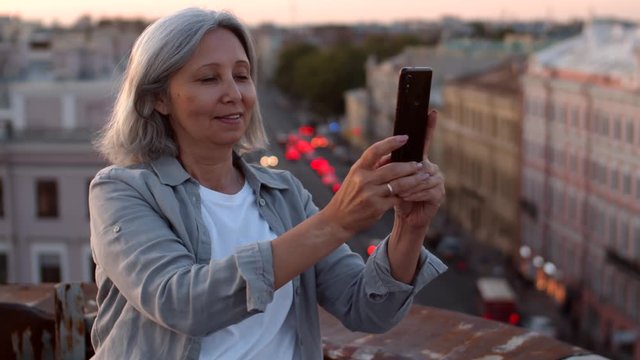 Medium shot of mixed-race attractive woman with grey hair standing alone on roof in historical center of city with smartphone in her hands and making photos of cityscape