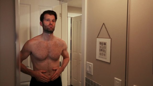 Handsome young caucasian man looking at himself in a bathroom mirror while examining his belly fat. - HD 24FPS.