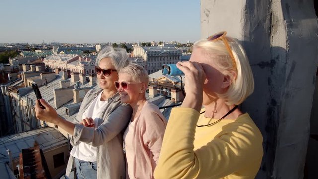 Medium shot of three diverse middle-aged women standing on roof in historical city center. Two women making photos on smartphone and one woman looking ahead through binoculars