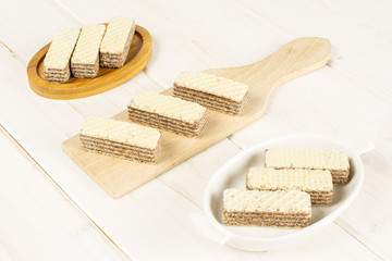 Group of nine whole crispy beige hazelnut wafer cookie in white oval ceramic bowl on bamboo coaster on small wooden cutting board on white wood