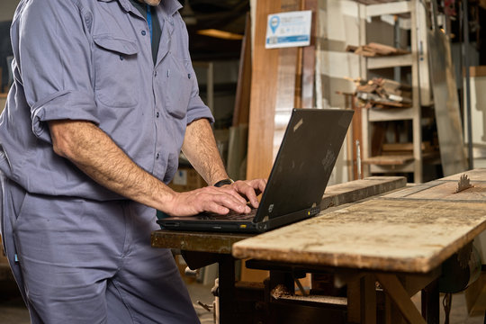 Man With Blue Overalls In Carpentry Using Computer In Workshop. Internet Communication Concept