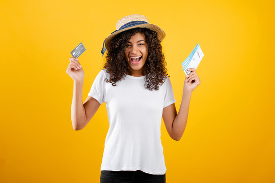 Smiling Happy Black Woman With Plane Tickets And Credit Card Wearing Hat Isolated Over Yellow