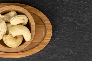 Lot of whole brown nut cashew on round bamboo coaster in bamboo bowl flatlay on grey stone